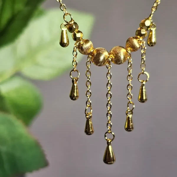 Elegante Goldene Tropfen-Fransen Halskette – Vintage Laser-Design mit schimmernden Kugeln Goldfarbene Modeschmuck-Halskette aus Kupfer, bestehend aus einer feinen Kette und einem mittigen Anhänger mit drei geriffelten Laser-Kugeln, von denen fünf filigrane Ketten mit kleinen Tropfen-Anhängern herabfallen.