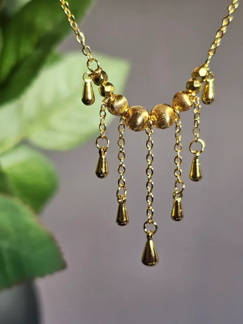 Elegante Goldene Tropfen-Fransen Halskette – Vintage Laser-Design mit schimmernden Kugeln Goldfarbene Modeschmuck-Halskette aus Kupfer, bestehend aus einer feinen Kette und einem mittigen Anhänger mit drei geriffelten Laser-Kugeln, von denen fünf filigrane Ketten mit kleinen Tropfen-Anhängern herabfallen.