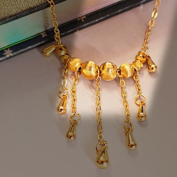 Elegante Goldene Tropfen-Fransen Halskette – Vintage Laser-Design mit schimmernden Kugeln Goldfarbene Modeschmuck-Halskette aus Kupfer, bestehend aus einer feinen Kette und einem mittigen Anhänger mit drei geriffelten Laser-Kugeln, von denen fünf filigrane Ketten mit kleinen Tropfen-Anhängern herabfallen.