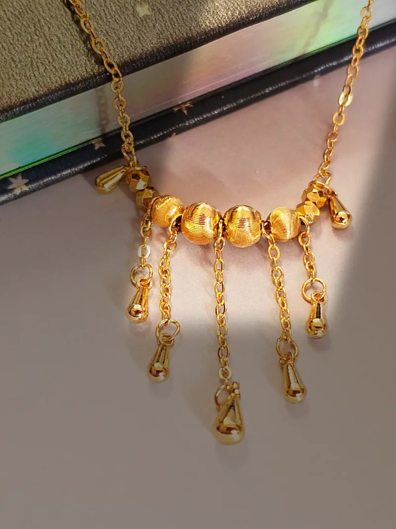 Elegante Goldene Tropfen-Fransen Halskette – Vintage Laser-Design mit schimmernden Kugeln Goldfarbene Modeschmuck-Halskette aus Kupfer, bestehend aus einer feinen Kette und einem mittigen Anhänger mit drei geriffelten Laser-Kugeln, von denen fünf filigrane Ketten mit kleinen Tropfen-Anhängern herabfallen.