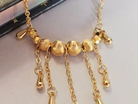 Elegante Goldene Tropfen-Fransen Halskette – Vintage Laser-Design mit schimmernden Kugeln Goldfarbene Modeschmuck-Halskette aus Kupfer, bestehend aus einer feinen Kette und einem mittigen Anhänger mit drei geriffelten Laser-Kugeln, von denen fünf filigrane Ketten mit kleinen Tropfen-Anhängern herabfallen.