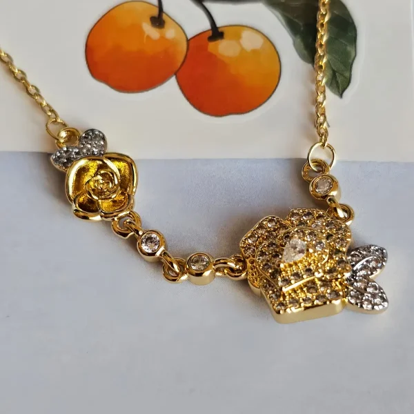Luxuriöse Rosengold-Halskette mit Kristallbesatz – Filigranes Doppel-Blüten Design Nahaufnahme einer goldfarbenen Halskette aus Kupfer. Der Anhänger zeigt zwei stilisierte Rosenblüten: Eine Blüte ist glatt und poliert, die andere ist dicht mit kleinen, funkelnden Kristallen (Pavé-Optik) besetzt und hat einen größeren Kristall in der Mitte. Die Kette verbindet die Elemente mit kleinen, runden Kristallsteinen.