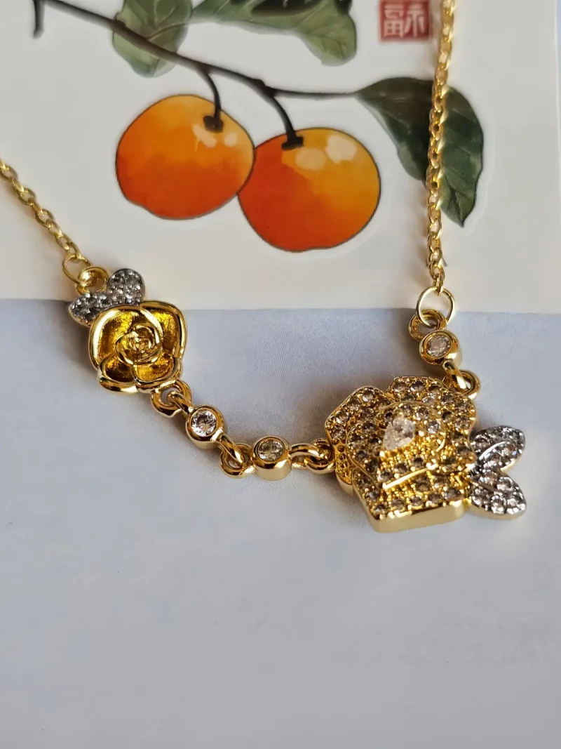 Luxuriöse Rosengold-Halskette mit Kristallbesatz – Filigranes Doppel-Blüten Design Nahaufnahme einer goldfarbenen Halskette aus Kupfer. Der Anhänger zeigt zwei stilisierte Rosenblüten: Eine Blüte ist glatt und poliert, die andere ist dicht mit kleinen, funkelnden Kristallen (Pavé-Optik) besetzt und hat einen größeren Kristall in der Mitte. Die Kette verbindet die Elemente mit kleinen, runden Kristallsteinen.