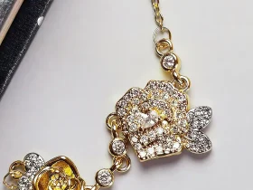 Luxuriöse Rosengold-Halskette mit Kristallbesatz – Filigranes Doppel-Blüten Design Nahaufnahme einer goldfarbenen Halskette aus Kupfer. Der Anhänger zeigt zwei stilisierte Rosenblüten: Eine Blüte ist glatt und poliert, die andere ist dicht mit kleinen, funkelnden Kristallen (Pavé-Optik) besetzt und hat einen größeren Kristall in der Mitte. Die Kette verbindet die Elemente mit kleinen, runden Kristallsteinen.