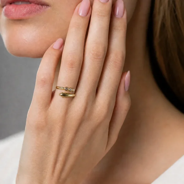 Nahaufnahme einer Frauenhand mit einem goldenen Schlangenring, der mit drei kleinen Zirkonia-Steinen besetzt ist. Der Ring wird elegant am Kinn präsentiert.