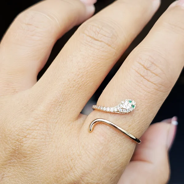 Eleganter, goldfarbener Schlangenring im offenen Design, mit funkelnden weißen Schmucksteinen und zwei grünen Augen. Nahaufnahme des Rings an einer Hand vor dunklem Hintergrund.