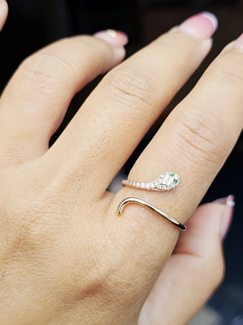 Eleganter, goldfarbener Schlangenring im offenen Design, mit funkelnden weißen Schmucksteinen und zwei grünen Augen. Nahaufnahme des Rings an einer Hand vor dunklem Hintergrund.
