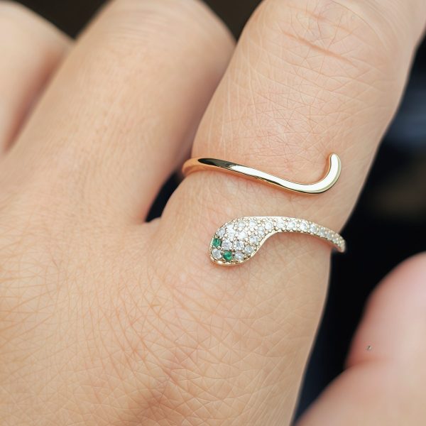 Eleganter, goldfarbener Schlangenring im offenen Design, mit funkelnden weißen Schmucksteinen und zwei grünen Augen. Nahaufnahme des Rings an einer Hand vor dunklem Hintergrund.