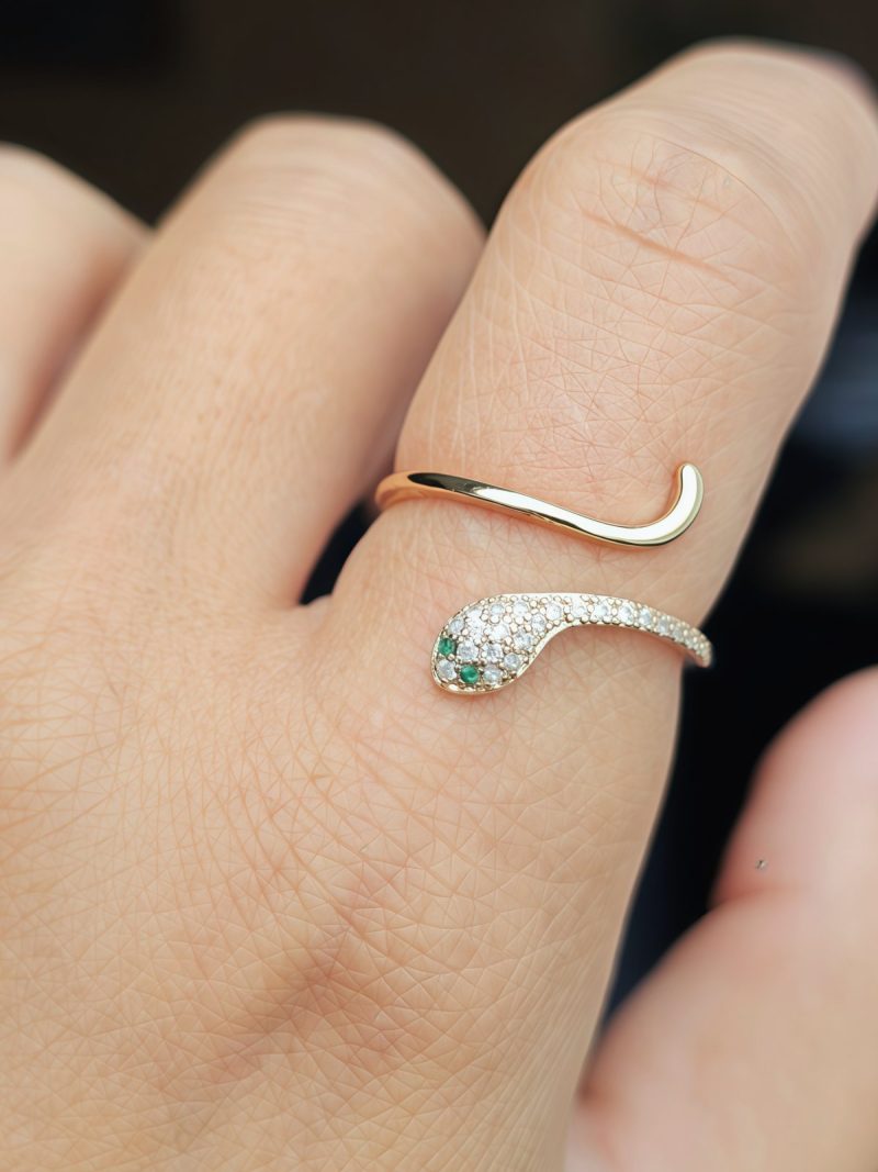 Eleganter, goldfarbener Schlangenring im offenen Design, mit funkelnden weißen Schmucksteinen und zwei grünen Augen. Nahaufnahme des Rings an einer Hand vor dunklem Hintergrund.