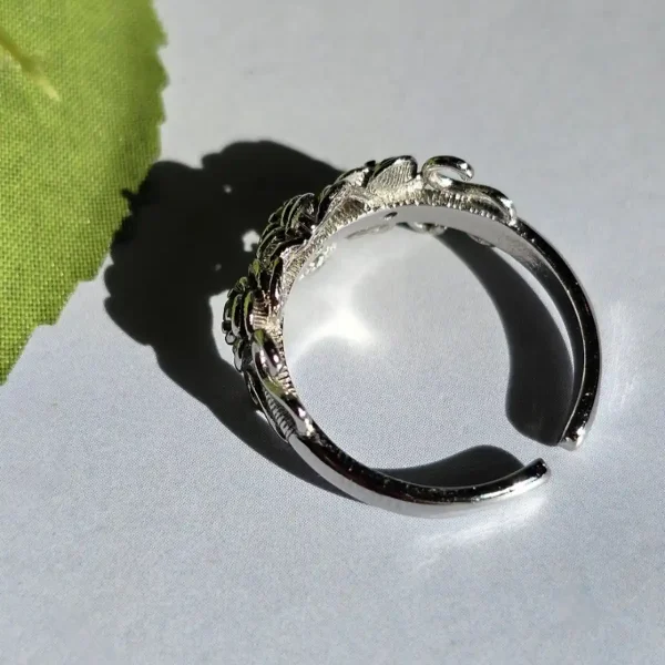 Vintage Rose Ring aus Weißkupfer – Statement-Schmuck mit Zirkonia für sie Ein offener silberfarbener Ring mit Rosen-Design und filigranen Details, seitlich auf einer weißen Oberfläche neben einem grünen Blatt platziert.