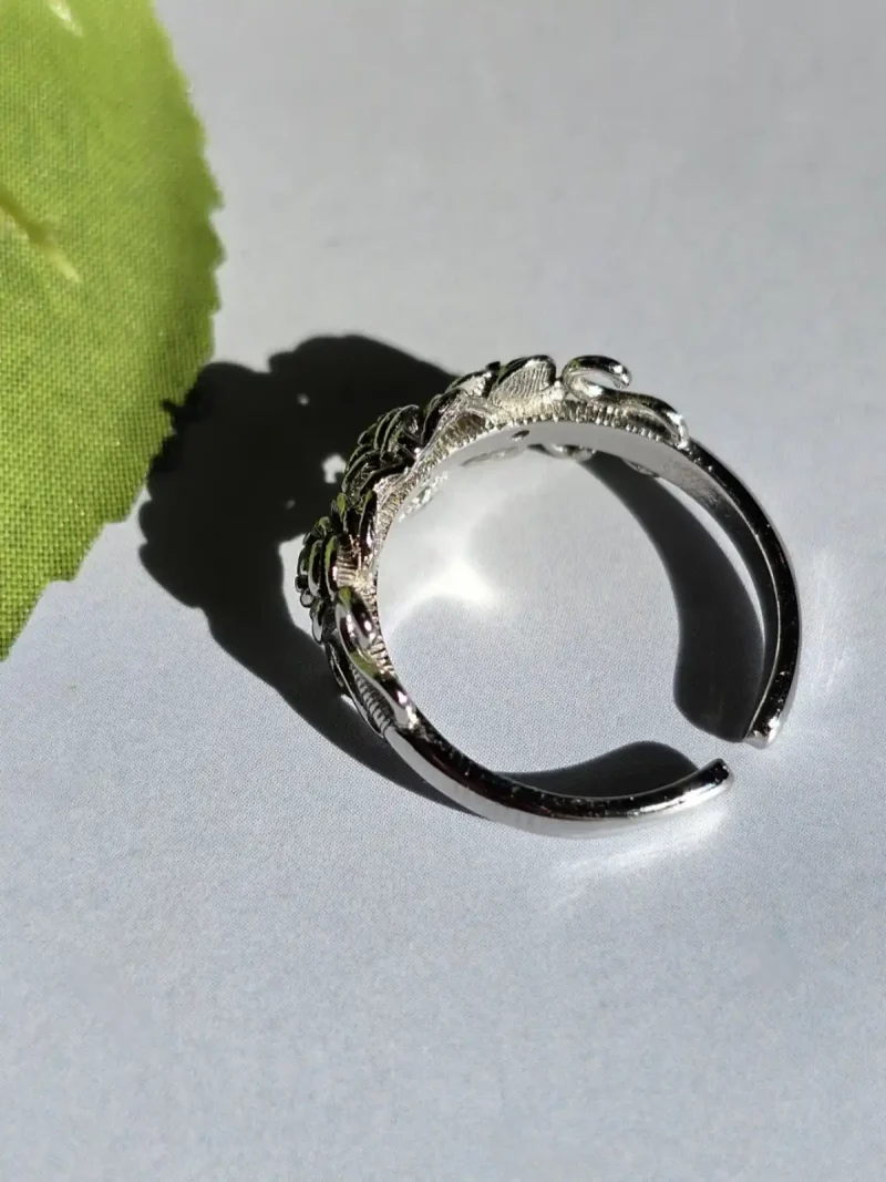 Vintage Rose Ring aus Weißkupfer – Statement-Schmuck mit Zirkonia für sie Ein offener silberfarbener Ring mit Rosen-Design und filigranen Details, seitlich auf einer weißen Oberfläche neben einem grünen Blatt platziert.