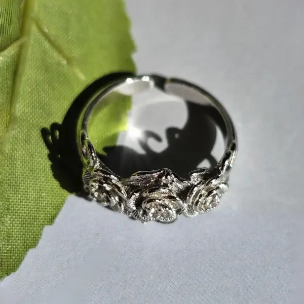 Vintage Rose Ring aus Weißkupfer – Statement-Schmuck mit Zirkonia für sie Ein silberfarbener Ring im Vintage-Stil mit drei Rosen und Zirkonia-Steinen, liegend auf einem weißen Untergrund neben einem grünen Blatt.
