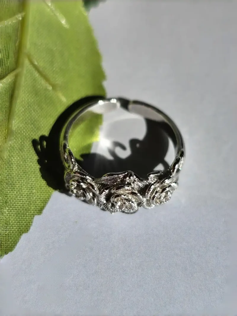 Vintage Rose Ring aus Weißkupfer – Statement-Schmuck mit Zirkonia für sie Ein silberfarbener Ring im Vintage-Stil mit drei Rosen und Zirkonia-Steinen, liegend auf einem weißen Untergrund neben einem grünen Blatt.