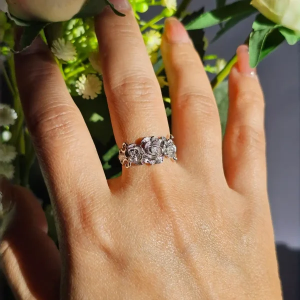 Vintage Rose Ring aus Weißkupfer – Statement-Schmuck mit Zirkonia für sie Eine Hand, die einen silberfarbenen Vintage-Ring mit Rosen-Design und Zirkonia-Steinen trägt, umgeben von weißen Rosen und grünen Blumen im Hintergrund.