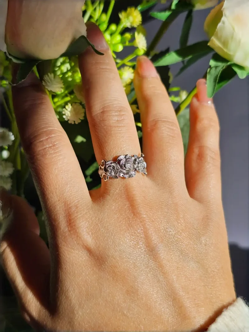 Vintage Rose Ring aus Weißkupfer – Statement-Schmuck mit Zirkonia für sie Eine Hand, die einen silberfarbenen Vintage-Ring mit Rosen-Design und Zirkonia-Steinen trägt, umgeben von weißen Rosen und grünen Blumen im Hintergrund.