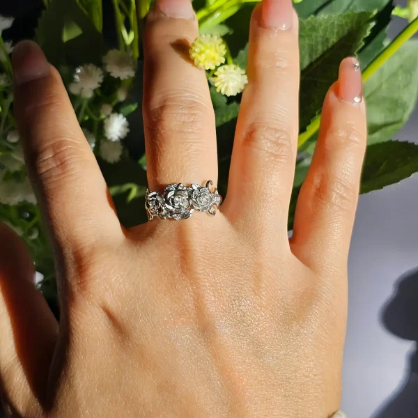 Vintage Rose Ring aus Weißkupfer – Statement-Schmuck mit Zirkonia für sie Eine Hand, die einen silberfarbenen Vintage-Ring mit Rosen-Design und Zirkonia-Steinen trägt, umgeben von weißen Rosen und grünen Blumen im Hintergrund.