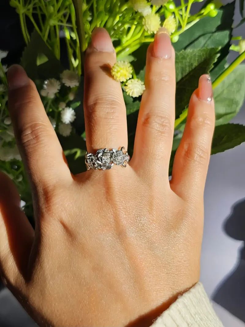 Vintage Rose Ring aus Weißkupfer – Statement-Schmuck mit Zirkonia für sie Eine Hand, die einen silberfarbenen Vintage-Ring mit Rosen-Design und Zirkonia-Steinen trägt, umgeben von weißen Rosen und grünen Blumen im Hintergrund.