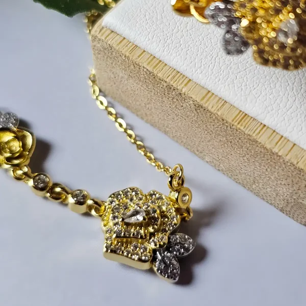 Schmuckset mit Halskette und Damenring in Rosen Design Nahaufnahme einer goldfarbenen Geschenke Schmuckset-Halskette. Der Set zeigt zwei stilisierte Rosenblüten: Eine Blüte ist glatt und poliert, die andere ist dicht mit kleinen, funkelnden Kristallen (Pavé-Optik) besetzt und hat einen größeren Kristall in der Mitte. Die Kette verbindet die Elemente mit kleinen, runden Kristallsteinen.