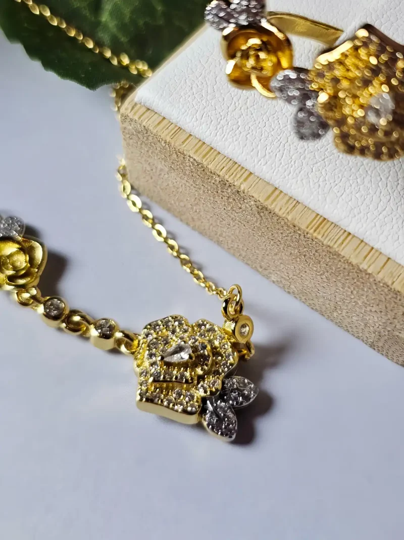 Schmuckset mit Halskette und Damenring in Rosen Design Nahaufnahme einer goldfarbenen Geschenke Schmuckset-Halskette. Der Set zeigt zwei stilisierte Rosenblüten: Eine Blüte ist glatt und poliert, die andere ist dicht mit kleinen, funkelnden Kristallen (Pavé-Optik) besetzt und hat einen größeren Kristall in der Mitte. Die Kette verbindet die Elemente mit kleinen, runden Kristallsteinen.