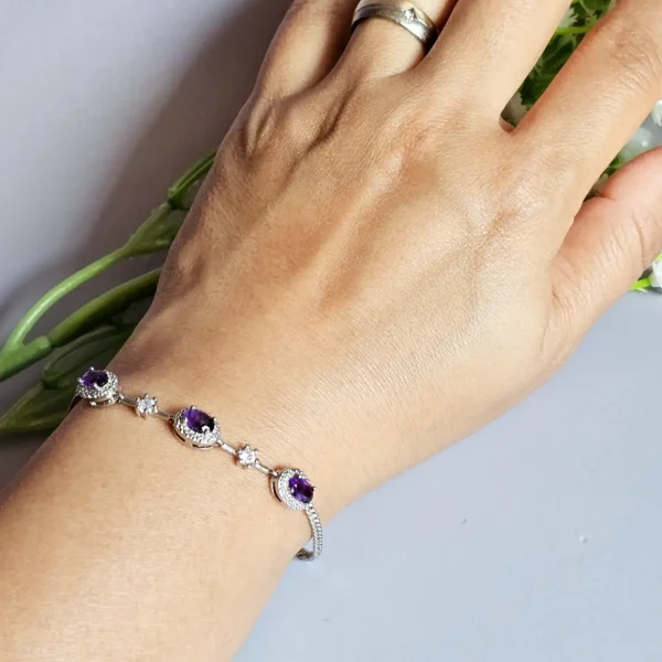 Amethyst Armband aus Kupfer versilbert Nahaufnahme eines Handgelenks mit einem eleganten Silberarmband, das mit drei violetten Edelsteinen und funkelnden Schmucksteinen verziert ist. Im Hintergrund sind grüne Stiele und kleine weiße Blüten zu sehen.
