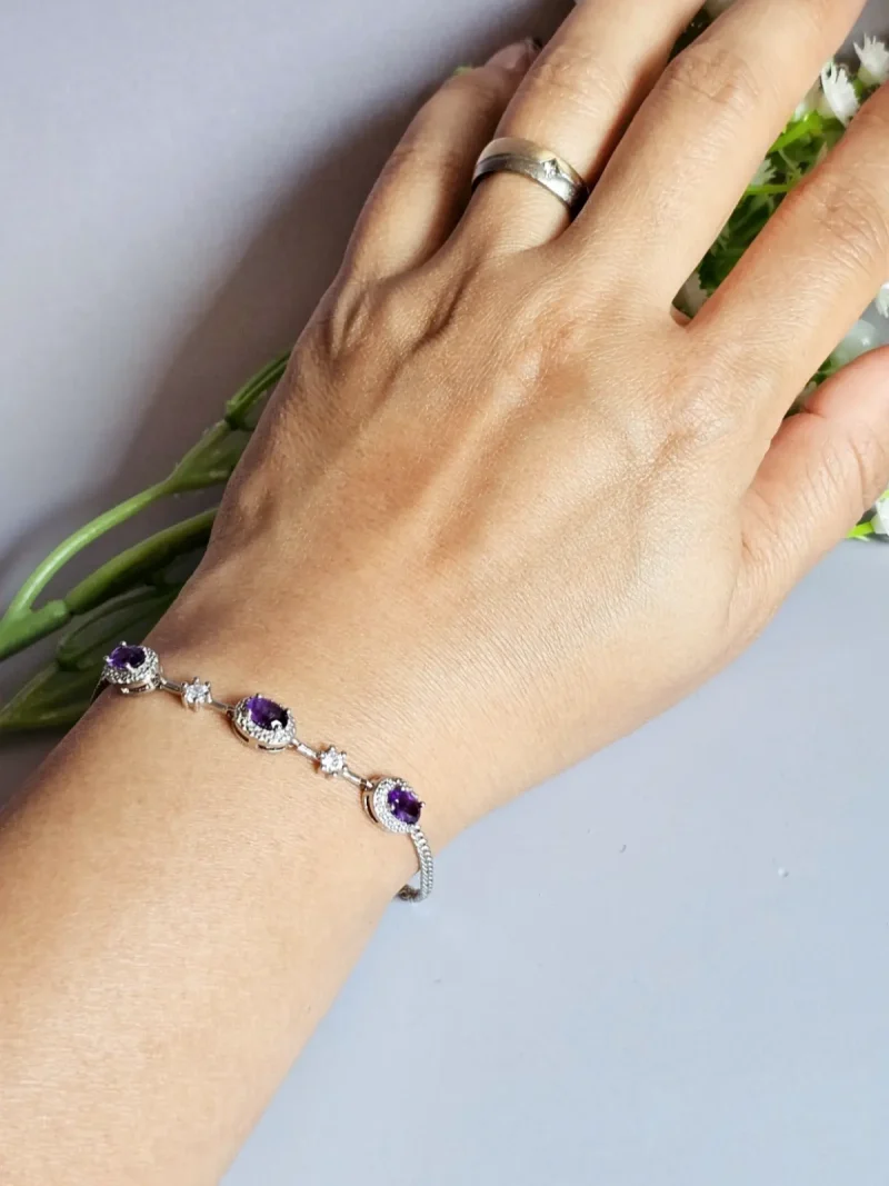 Amethyst Armband aus Kupfer versilbert Nahaufnahme eines Handgelenks mit einem eleganten Silberarmband, das mit drei violetten Edelsteinen und funkelnden Schmucksteinen verziert ist. Im Hintergrund sind grüne Stiele und kleine weiße Blüten zu sehen.