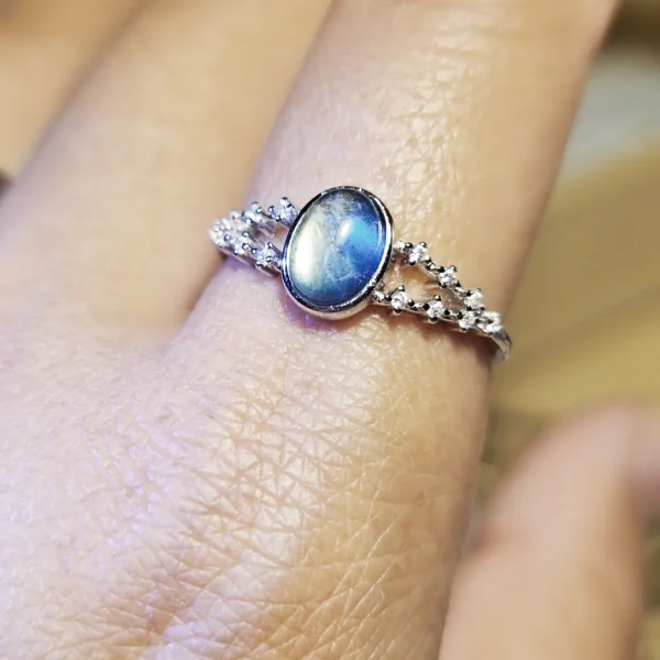 DR-SIL-03-06 Silberring mit ovalem, blau schimmerndem Mondstein und funkelnden Zirkonia-Verzierungen, am Finger getragen.