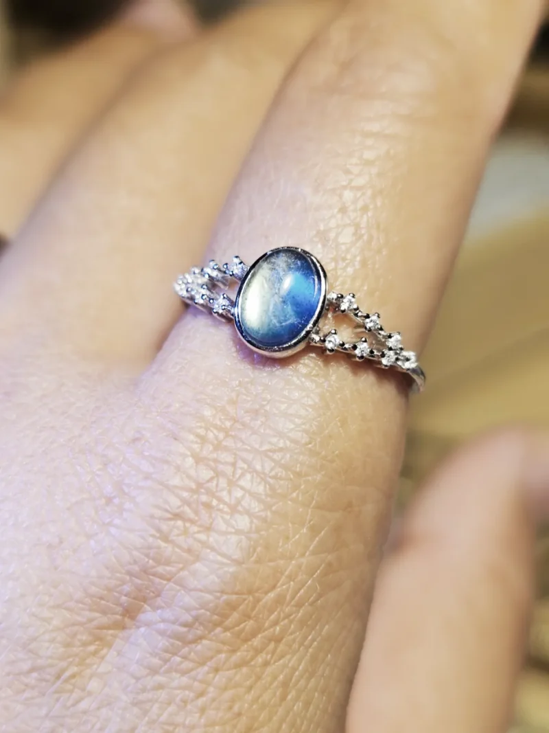 DR-SIL-03-06 Silberring mit ovalem, blau schimmerndem Mondstein und funkelnden Zirkonia-Verzierungen, am Finger getragen.