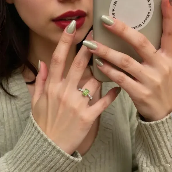 DR-SIL-03-4 Eine Frau mit olivgrünem Nagellack und rotem Lippenstift trägt einen 925er Silber-Ring mit rechteckigem Peridot und Zirkonia-Steinen am Ringfinger. Sie hält ein Smartphone mit einem runden Aufkleber und macht ein Selfie im Spiegel, während sie einen hellgrünen Strickpullover trägt.