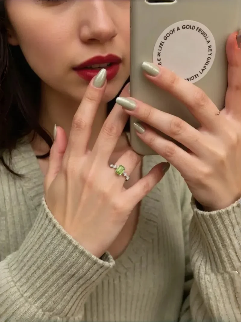 DR-SIL-03-4 Eine Frau mit olivgrünem Nagellack und rotem Lippenstift trägt einen 925er Silber-Ring mit rechteckigem Peridot und Zirkonia-Steinen am Ringfinger. Sie hält ein Smartphone mit einem runden Aufkleber und macht ein Selfie im Spiegel, während sie einen hellgrünen Strickpullover trägt.