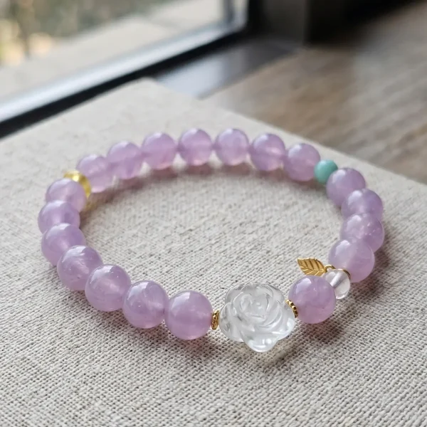 AB-PER-03-1 Ein elegantes Naturstein-Armband aus zartvioletten Amethystperlen mit einer geschnitzten Bergkristall-Rose, einer grünen Amazonitperle und vergoldeten Silber-Accessoires. Ein kleiner goldfarbener Blattanhänger rundet das Design ab. Das Armband liegt auf einem rosafarbenen Hintergrund im Sonnenlicht, das die transparenten Perlen sanft zum Leuchten bringt.