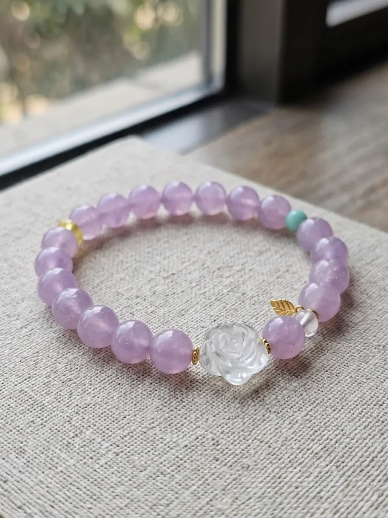 AB-PER-03-1 Ein elegantes Naturstein-Armband aus zartvioletten Amethystperlen mit einer geschnitzten Bergkristall-Rose, einer grünen Amazonitperle und vergoldeten Silber-Accessoires. Ein kleiner goldfarbener Blattanhänger rundet das Design ab. Das Armband liegt auf einem rosafarbenen Hintergrund im Sonnenlicht, das die transparenten Perlen sanft zum Leuchten bringt.