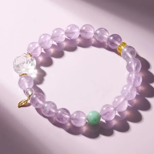 AB-PER-03-2 Ein elegantes Naturstein-Armband aus zartvioletten Amethystperlen mit einer geschnitzten Bergkristall-Rose, einer grünen Amazonitperle und vergoldeten Silber-Accessoires. Ein kleiner goldfarbener Blattanhänger rundet das Design ab. Das Armband liegt auf einem rosafarbenen Hintergrund im Sonnenlicht, das die transparenten Perlen sanft zum Leuchten bringt.