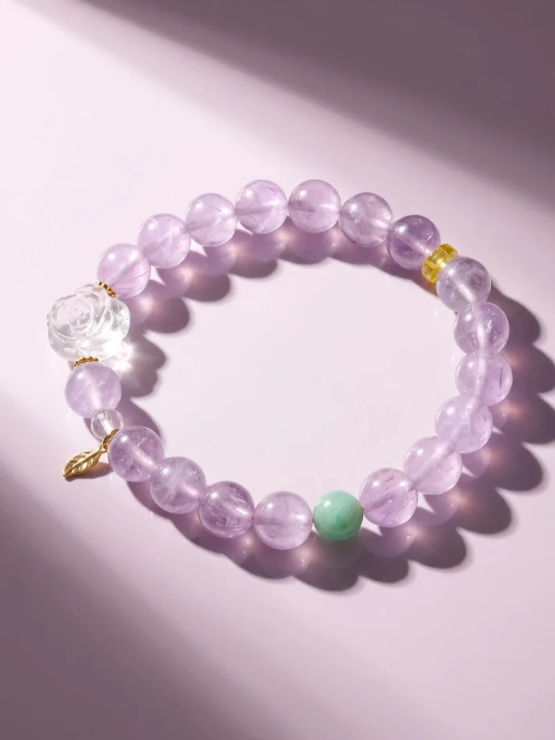AB-PER-03-2 Ein elegantes Naturstein-Armband aus zartvioletten Amethystperlen mit einer geschnitzten Bergkristall-Rose, einer grünen Amazonitperle und vergoldeten Silber-Accessoires. Ein kleiner goldfarbener Blattanhänger rundet das Design ab. Das Armband liegt auf einem rosafarbenen Hintergrund im Sonnenlicht, das die transparenten Perlen sanft zum Leuchten bringt.