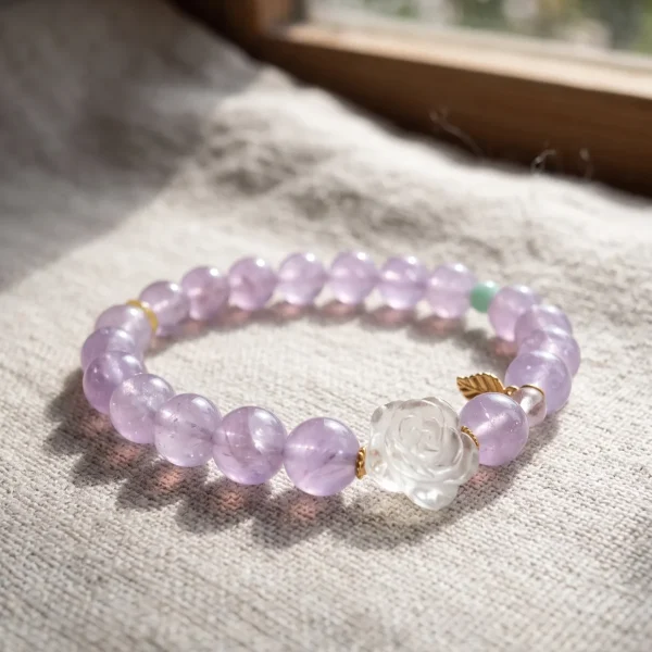 AB-PER-03-3 Ein elegantes Naturstein-Armband aus zartvioletten Amethystperlen mit einer geschnitzten Bergkristall-Rose, einer grünen Amazonitperle und vergoldeten Silber-Accessoires. Ein kleiner goldfarbener Blattanhänger rundet das Design ab. Das Armband liegt auf einem rosafarbenen Hintergrund im Sonnenlicht, das die transparenten Perlen sanft zum Leuchten bringt.