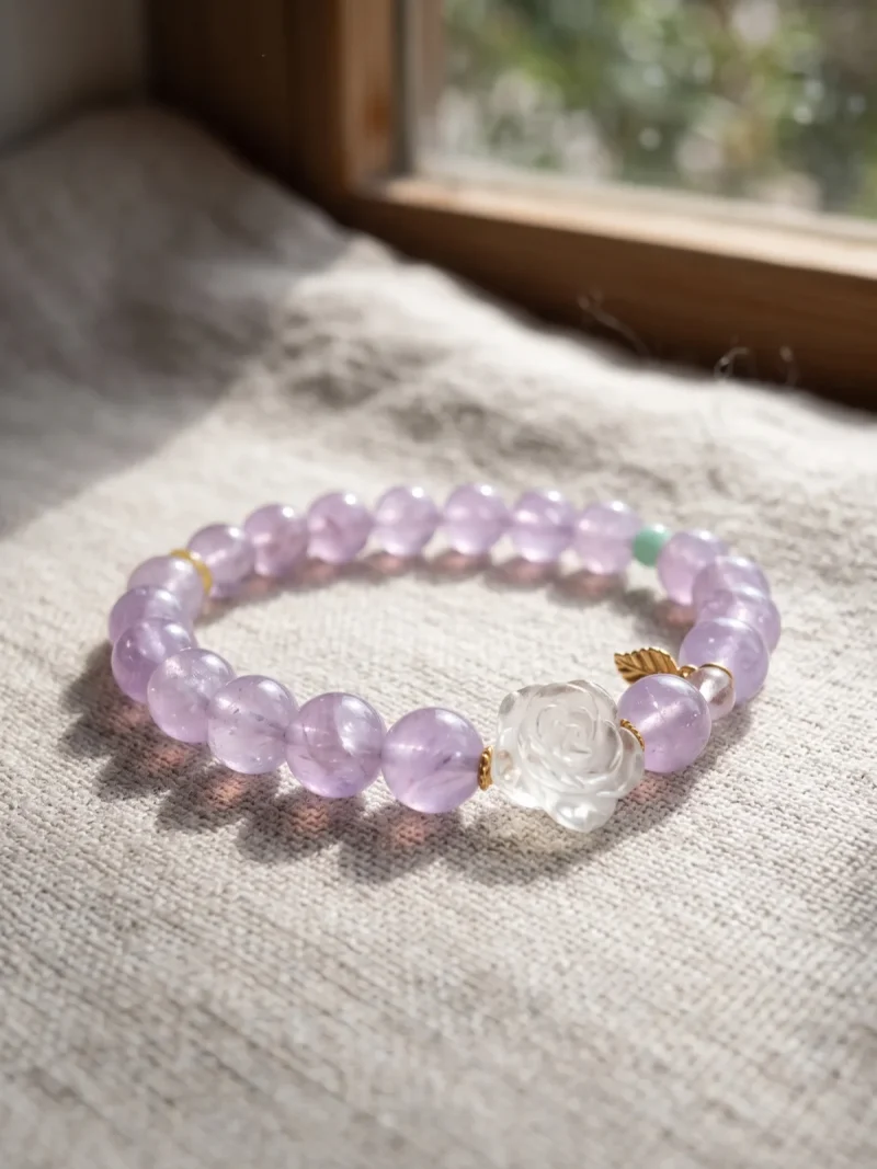 AB-PER-03-3 Ein elegantes Naturstein-Armband aus zartvioletten Amethystperlen mit einer geschnitzten Bergkristall-Rose, einer grünen Amazonitperle und vergoldeten Silber-Accessoires. Ein kleiner goldfarbener Blattanhänger rundet das Design ab. Das Armband liegt auf einem rosafarbenen Hintergrund im Sonnenlicht, das die transparenten Perlen sanft zum Leuchten bringt.