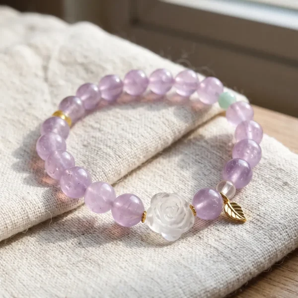 AB-PER-03-4 Ein elegantes Naturstein-Armband aus zartvioletten Amethystperlen mit einer geschnitzten Bergkristall-Rose, einer grünen Amazonitperle und vergoldeten Silber-Accessoires. Ein kleiner goldfarbener Blattanhänger rundet das Design ab. Das Armband liegt auf einem rosafarbenen Hintergrund im Sonnenlicht, das die transparenten Perlen sanft zum Leuchten bringt.