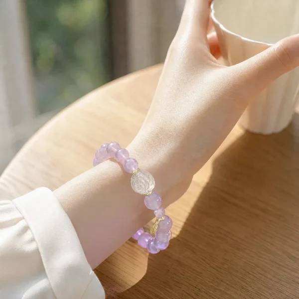 AB-PER-03-5 Eine elegante Nahaufnahme eines Handgelenks mit einem zartvioletten Amethyst-Armband. Die transparenten Natursteinperlen werden durch eine kunstvoll geschnitzte Bergkristall-Rose und kleine goldfarbene Details ergänzt. Warmes Sonnenlicht unterstreicht die sanften Lilatöne und verleiht dem Schmuck eine romantische, feminine Ausstrahlung.