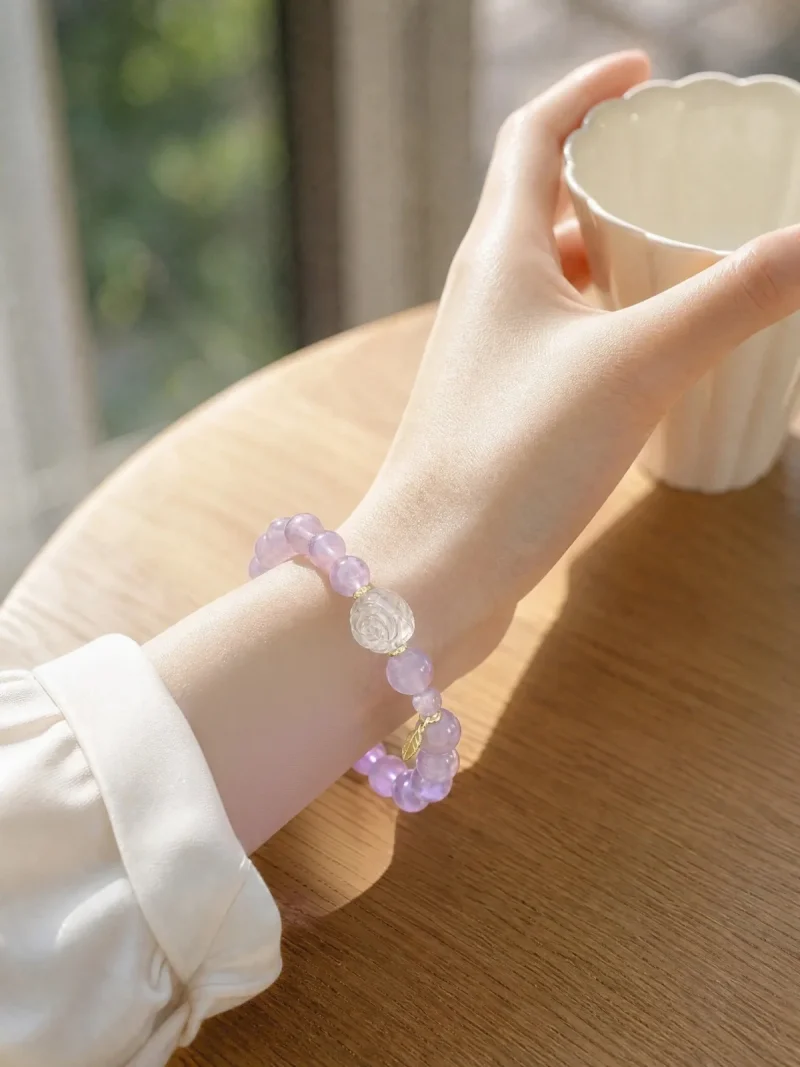 AB-PER-03-5 Eine elegante Nahaufnahme eines Handgelenks mit einem zartvioletten Amethyst-Armband. Die transparenten Natursteinperlen werden durch eine kunstvoll geschnitzte Bergkristall-Rose und kleine goldfarbene Details ergänzt. Warmes Sonnenlicht unterstreicht die sanften Lilatöne und verleiht dem Schmuck eine romantische, feminine Ausstrahlung.