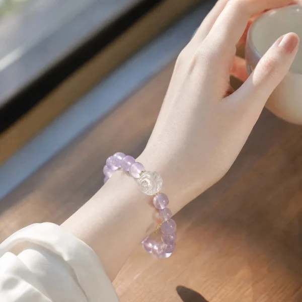 AB-PER-03-6 Eine elegante Nahaufnahme eines Handgelenks mit einem zartvioletten Amethyst-Armband. Die transparenten Natursteinperlen werden durch eine kunstvoll geschnitzte Bergkristall-Rose und kleine goldfarbene Details ergänzt. Warmes Sonnenlicht unterstreicht die sanften Lilatöne und verleiht dem Schmuck eine romantische, feminine Ausstrahlung.