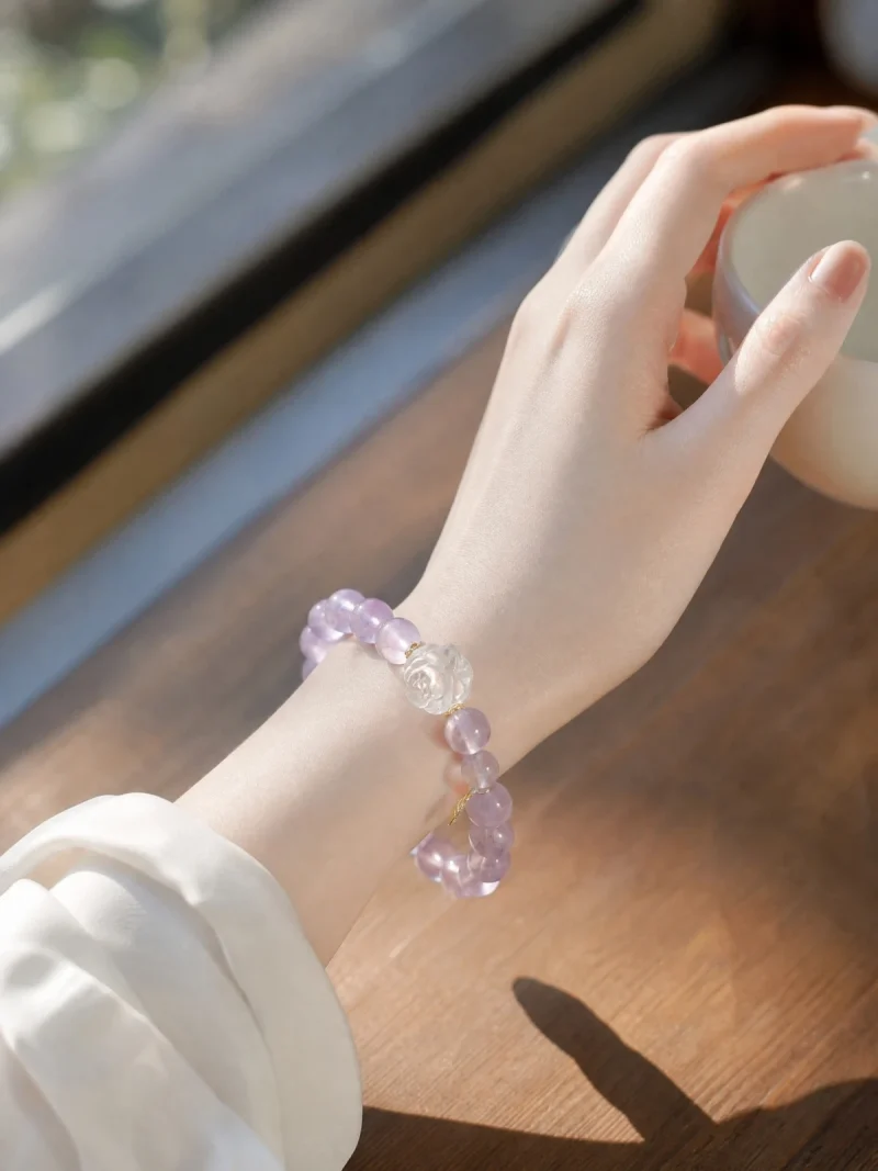 AB-PER-03-6 Eine elegante Nahaufnahme eines Handgelenks mit einem zartvioletten Amethyst-Armband. Die transparenten Natursteinperlen werden durch eine kunstvoll geschnitzte Bergkristall-Rose und kleine goldfarbene Details ergänzt. Warmes Sonnenlicht unterstreicht die sanften Lilatöne und verleiht dem Schmuck eine romantische, feminine Ausstrahlung.