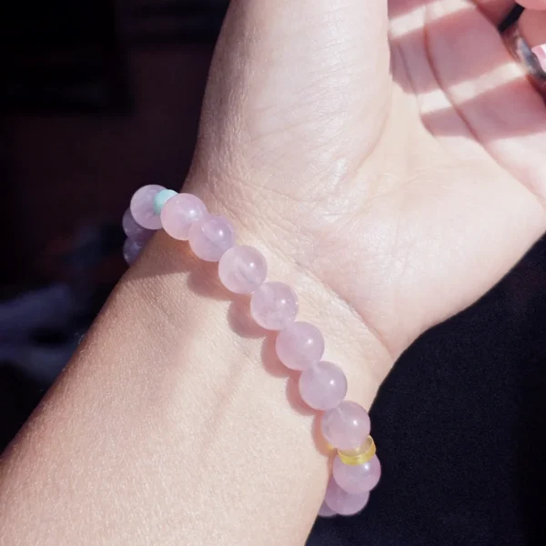 AB-PER-03-7 Eine elegante Nahaufnahme eines Handgelenks mit einem zartvioletten Amethyst-Armband. Die transparenten Natursteinperlen werden durch eine kunstvoll geschnitzte Bergkristall-Rose und kleine goldfarbene Details ergänzt. Warmes Sonnenlicht unterstreicht die sanften Lilatöne und verleiht dem Schmuck eine romantische, feminine Ausstrahlung.
