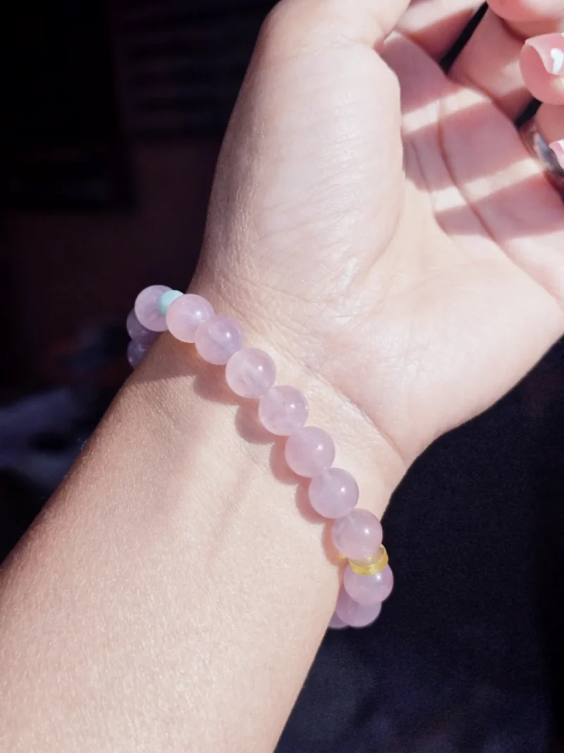 AB-PER-03-7 Eine elegante Nahaufnahme eines Handgelenks mit einem zartvioletten Amethyst-Armband. Die transparenten Natursteinperlen werden durch eine kunstvoll geschnitzte Bergkristall-Rose und kleine goldfarbene Details ergänzt. Warmes Sonnenlicht unterstreicht die sanften Lilatöne und verleiht dem Schmuck eine romantische, feminine Ausstrahlung.