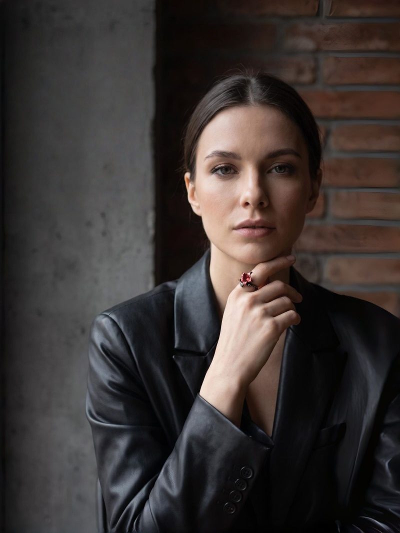 DR-GTC-01-9 Eine Frau trägt einen Gotischer Lilienring mit roter Blüte und gun-schwarzfarbener Vintage-Ringschiene im Dark Romantic Stil am Zeigfinger. Sie steht vor einem Wand.