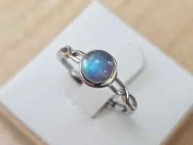 DR-TDS-09-01 Silberring mit rundem, blau schimmerndem Mondstein und filigraner, geschwungener Ringschiene auf weißem Schmuckdisplay.