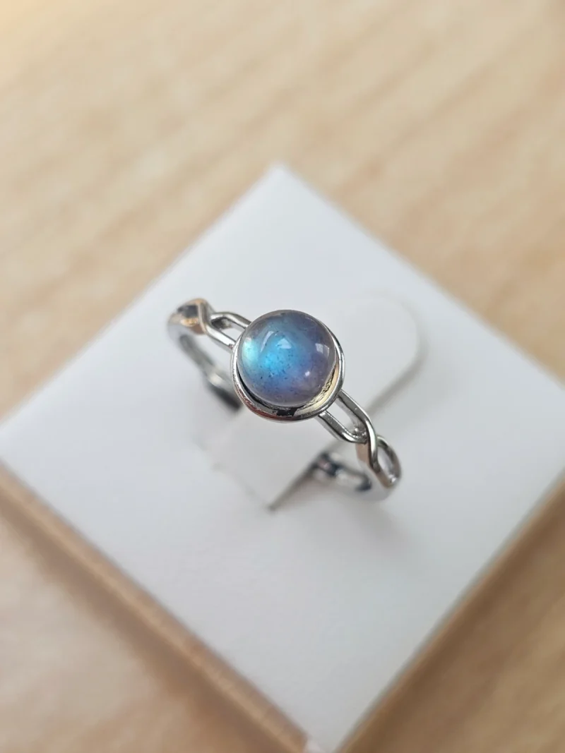 DR-TDS-09-01 Silberring mit rundem, blau schimmerndem Mondstein und filigraner, geschwungener Ringschiene auf weißem Schmuckdisplay.