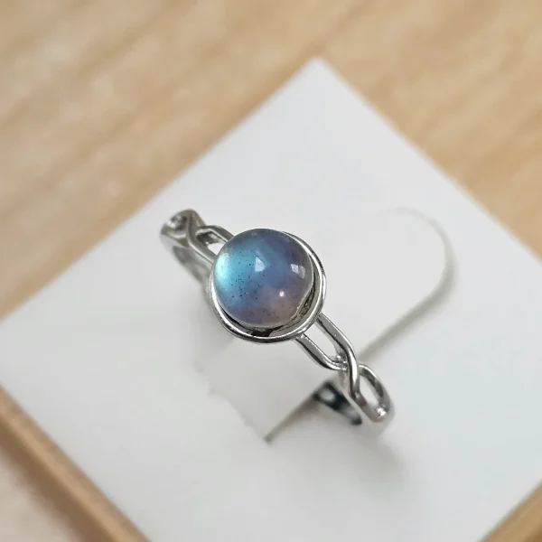DR-TDS-09-02 Silberring mit rundem, blau schimmerndem Mondstein und filigraner, geschwungener Ringschiene auf weißem Schmuckdisplay.