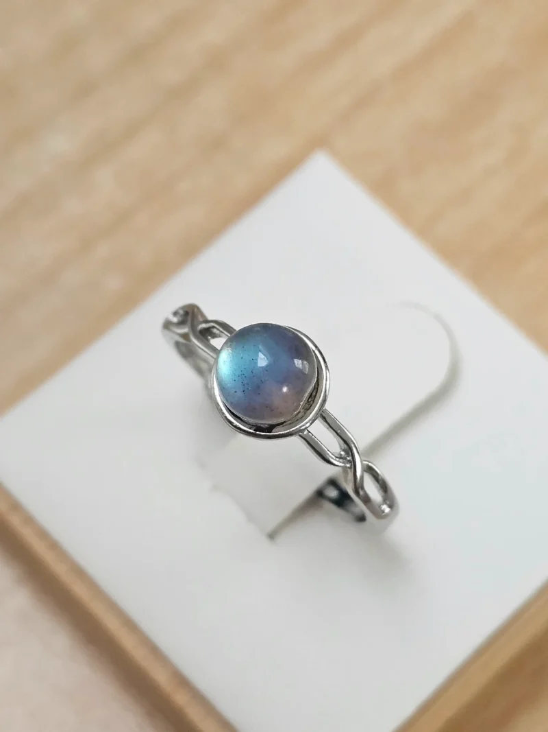 DR-TDS-09-02 Silberring mit rundem, blau schimmerndem Mondstein und filigraner, geschwungener Ringschiene auf weißem Schmuckdisplay.