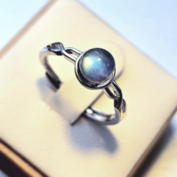 DR-TDS-09-03 Silberring mit rundem, blau schimmerndem Mondstein und filigraner, geschwungener Ringschiene auf weißem Schmuckdisplay.