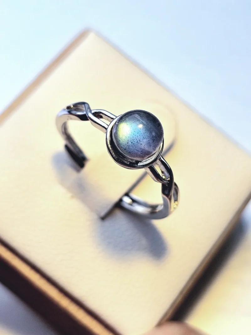 DR-TDS-09-03 Silberring mit rundem, blau schimmerndem Mondstein und filigraner, geschwungener Ringschiene auf weißem Schmuckdisplay.
